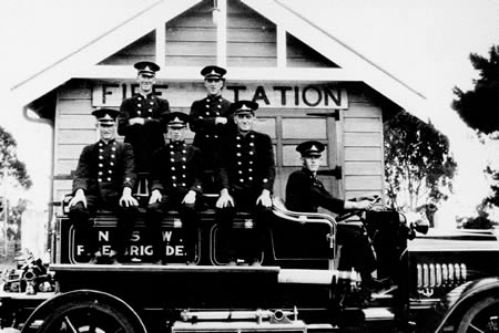 A black and white photo of fire fighters siting on a fire truck outside of their fire station.